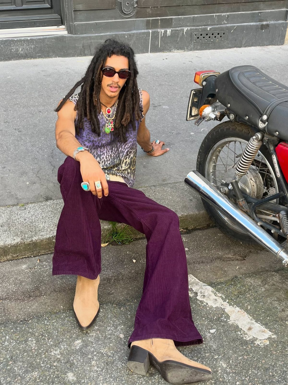 Man with dreadlocks wearing patterned top, purple corduroy trousers, and colorful jewelry sitting by a motorcycle.