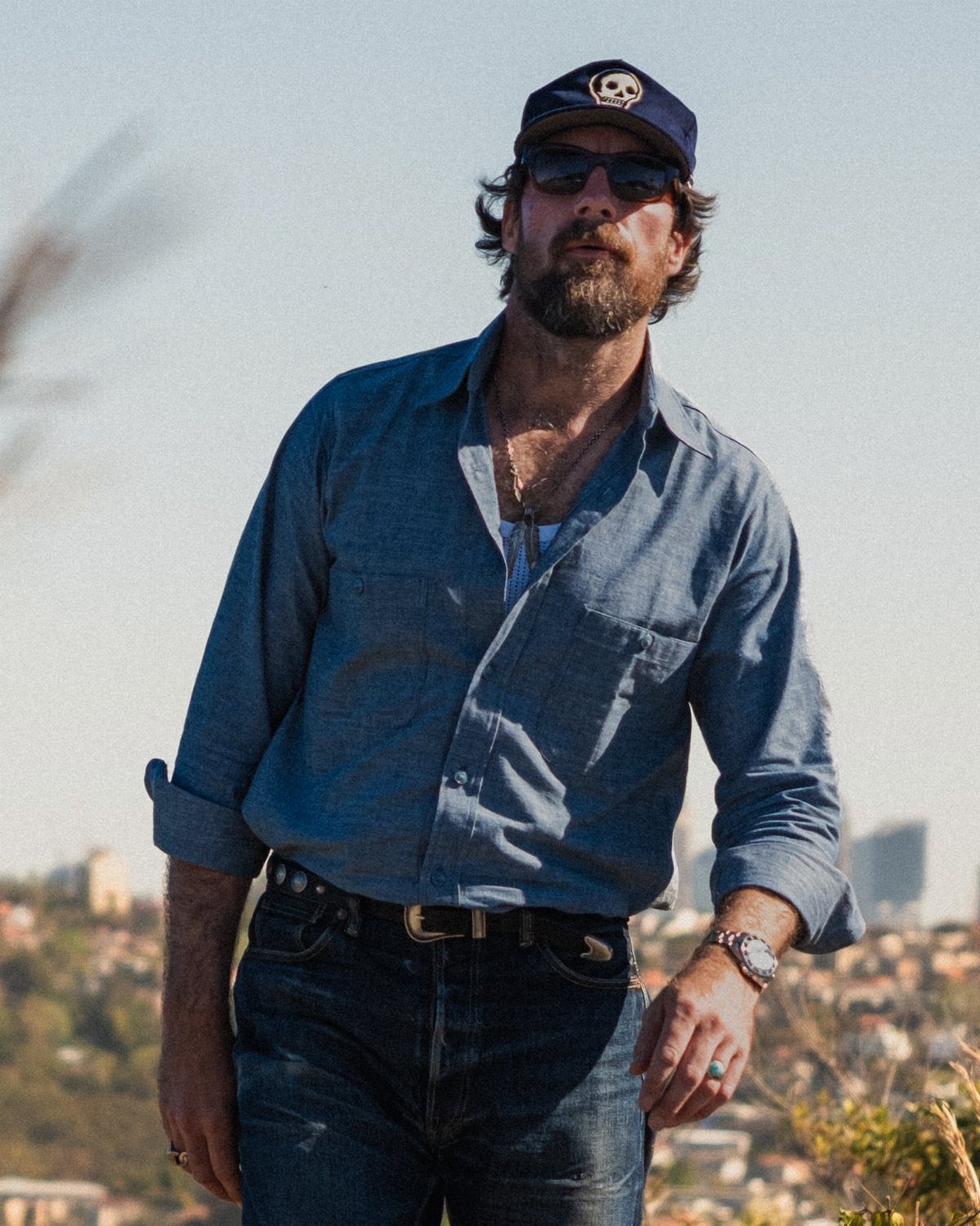A bearded man in a blue shirt, jeans, and a skull cap standing outdoors.