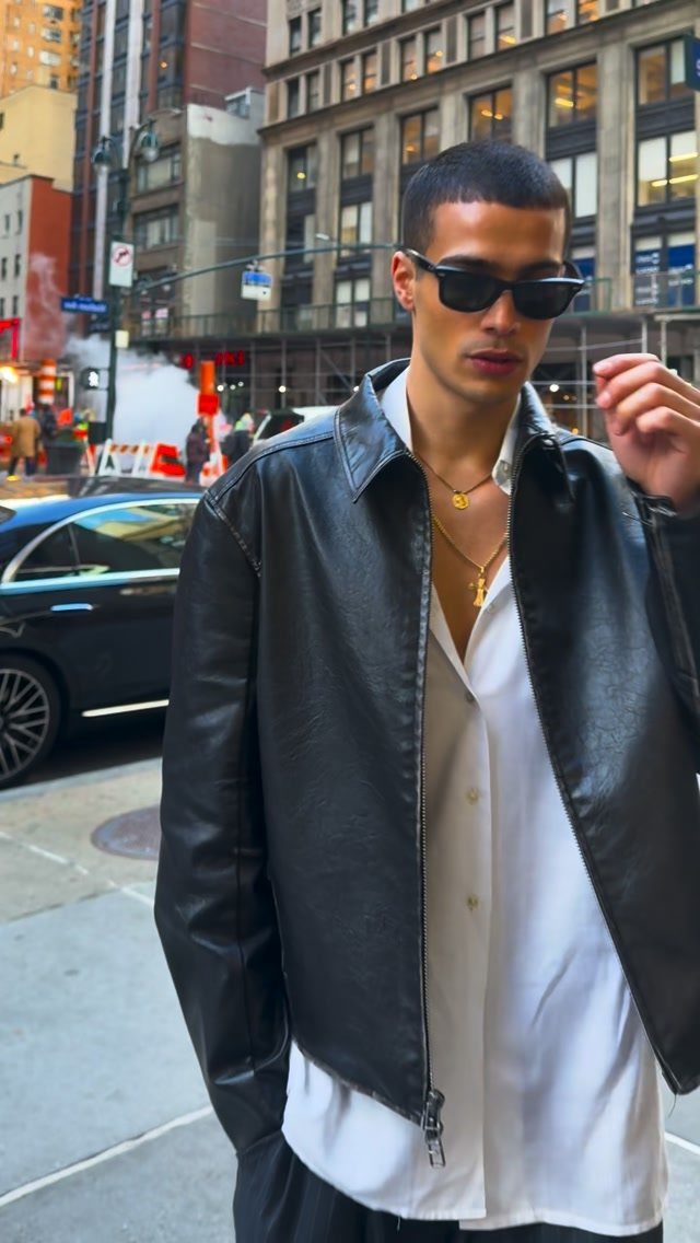 A man wearing a black leather jacket, white shirt, and sunglasses on a city street.