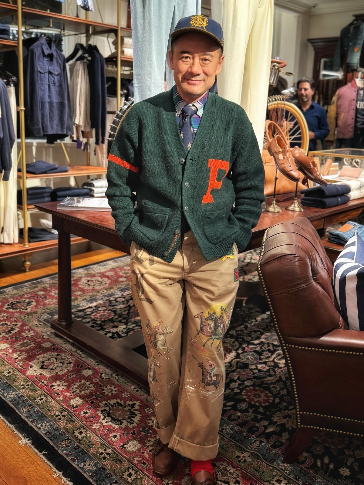 Man in a green cardigan, patterned tie, and embroidered tan trousers standing in a boutique.