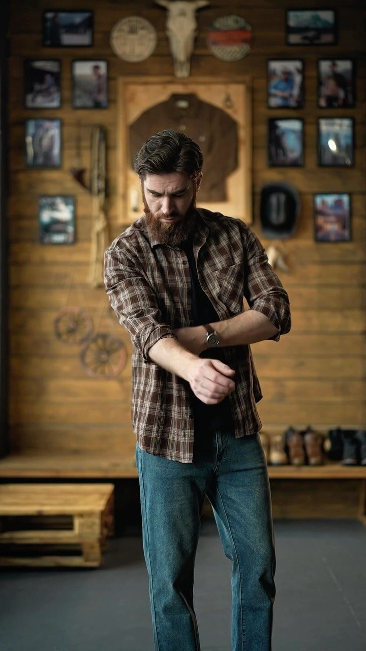 A bearded man in a plaid shirt and jeans standing in a rustic wooden room.