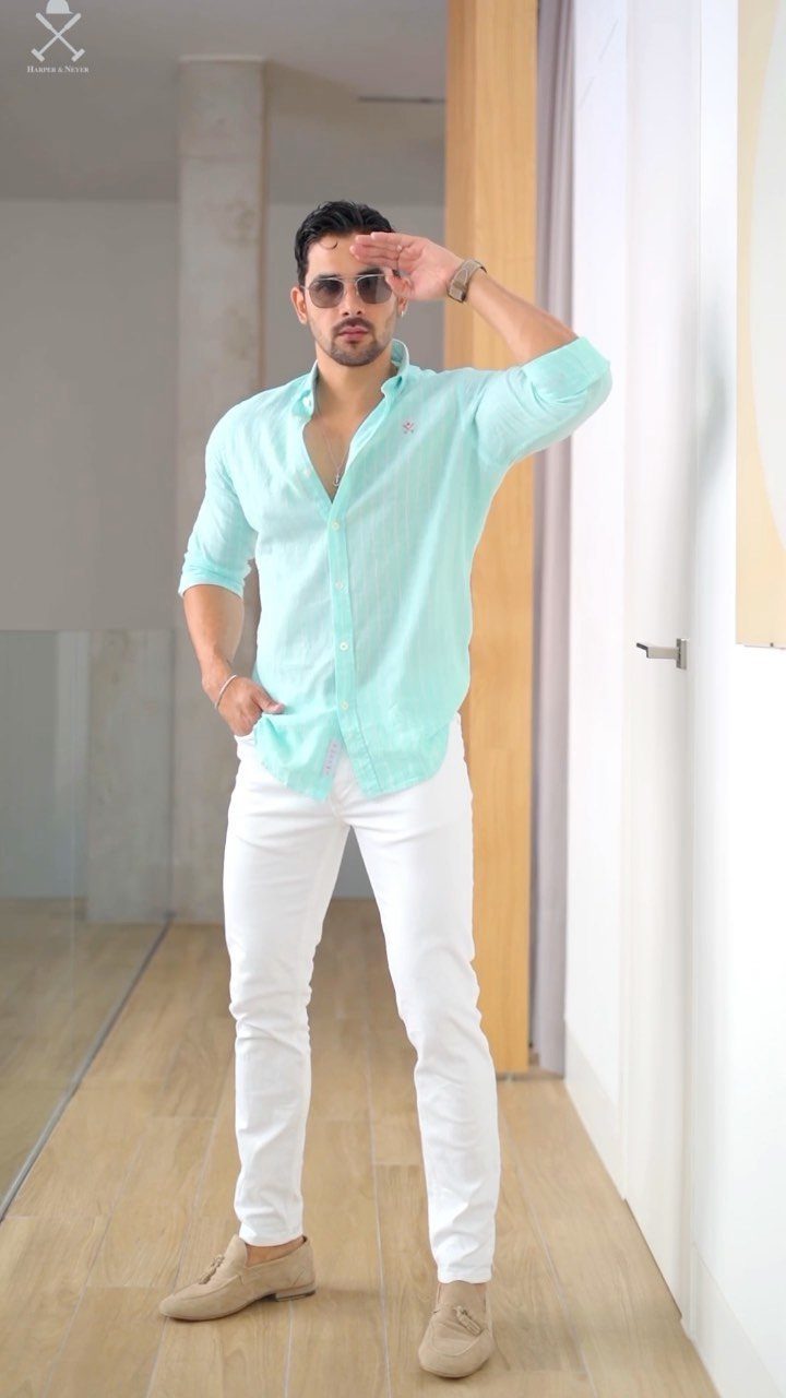 A man in a turquoise shirt and white pants posing in a bright hallway.