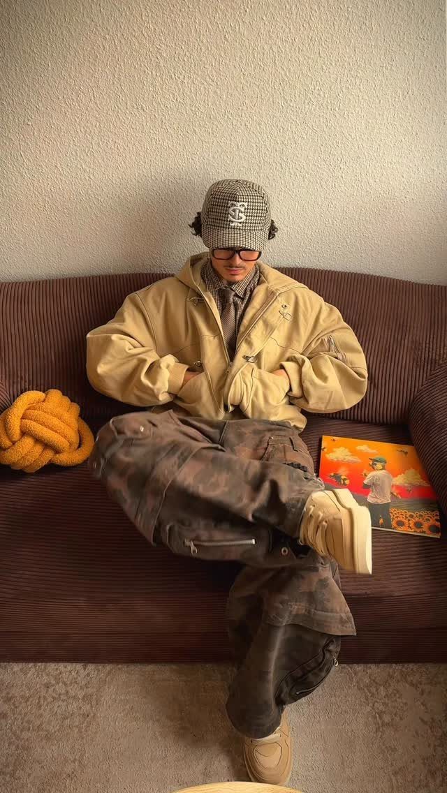 Man sitting on a brown sofa wearing a tan jacket and camouflage cargo pants.