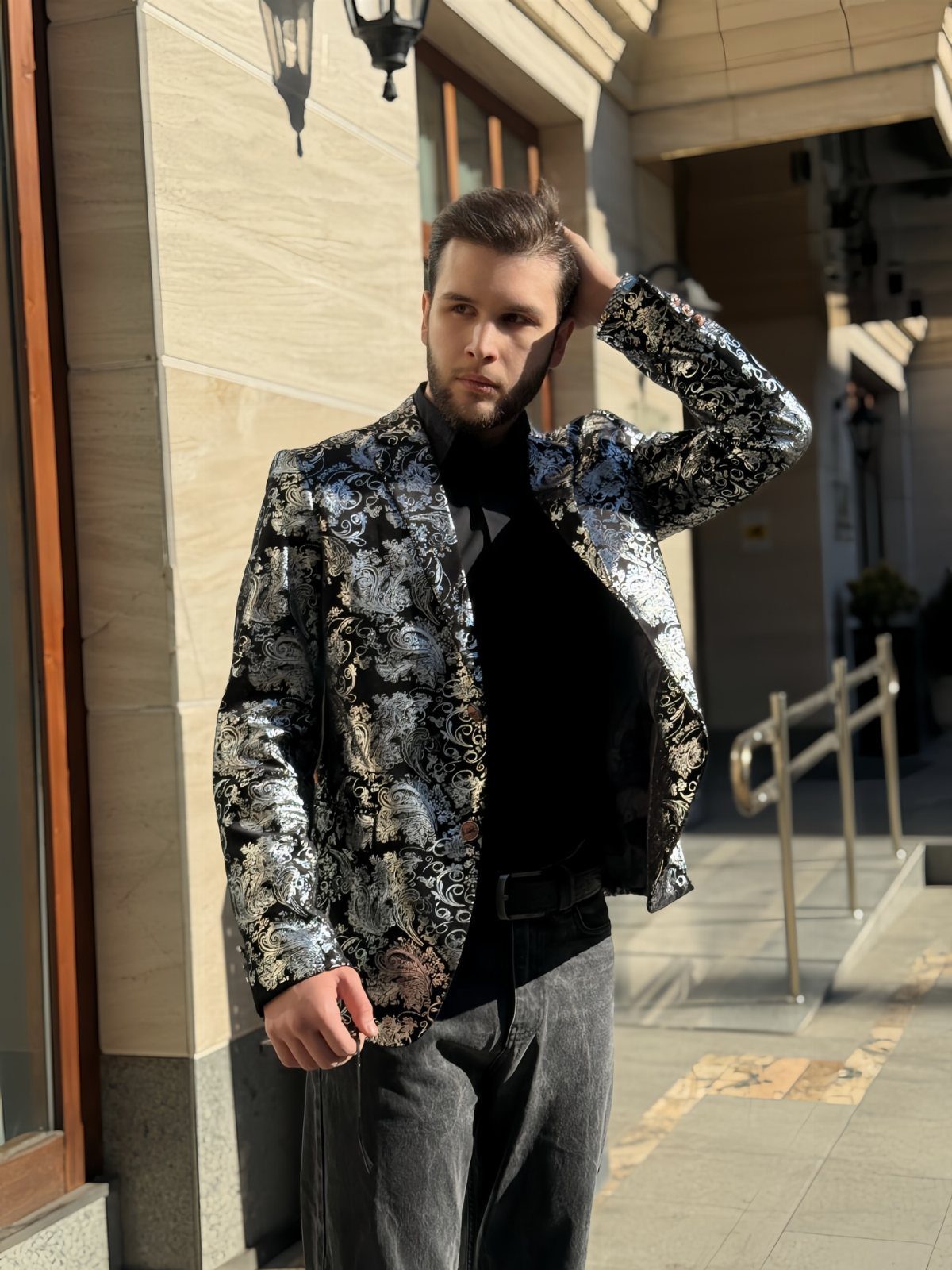 Man wearing a silver and black floral patterned blazer over a black turtleneck and dark trousers.