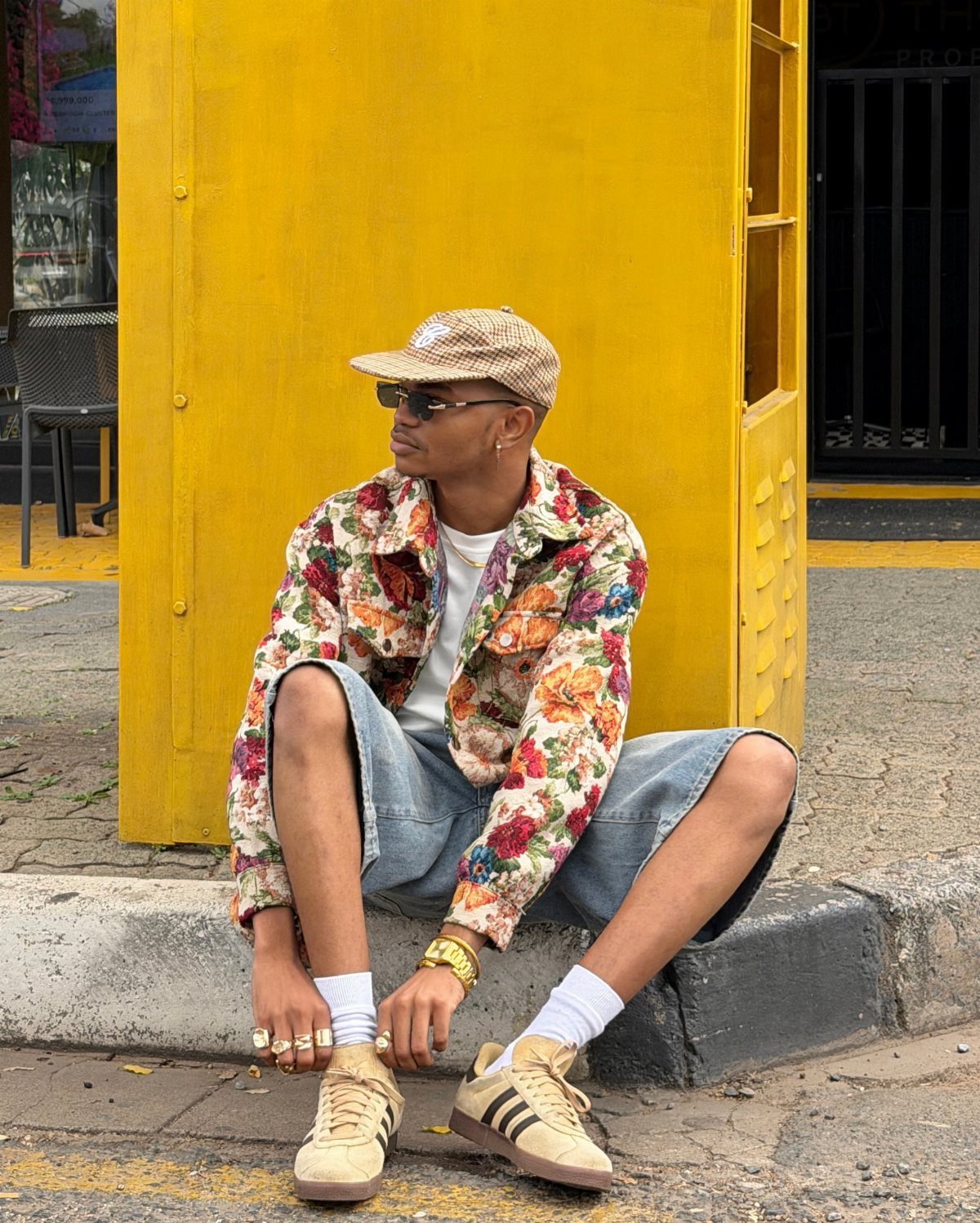 A man wearing a vibrant floral jacket and denim shorts sits against a yellow wall.