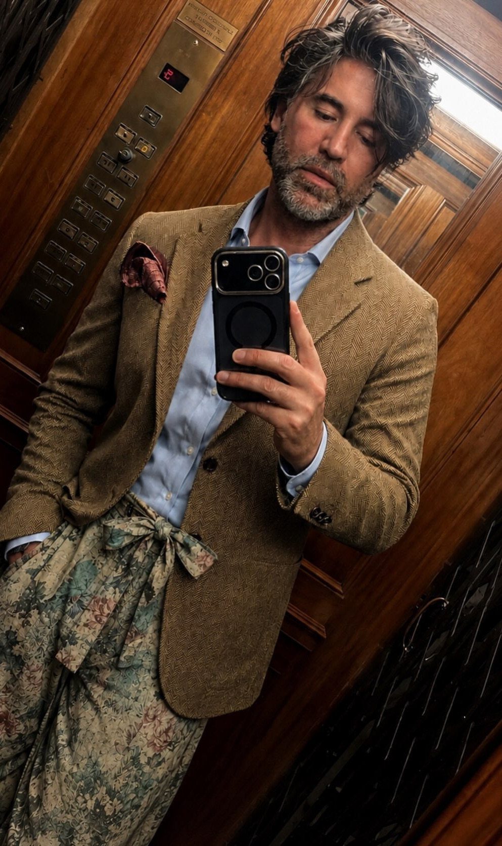 Man in a tan wool blazer, blue shirt, and floral trousers taking a selfie.