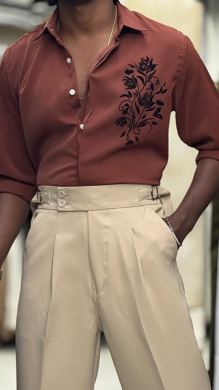 Man wearing a terracotta floral embroidered shirt tucked into beige pleated trousers.