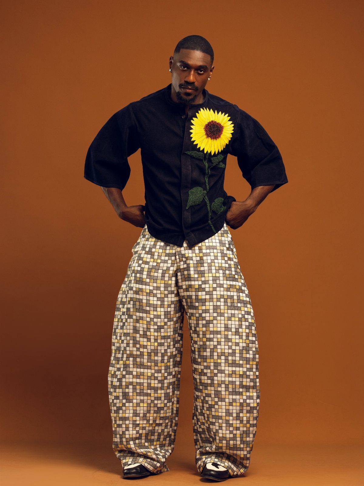 A man wearing a black shirt with a large sunflower and patterned wide-leg trousers.