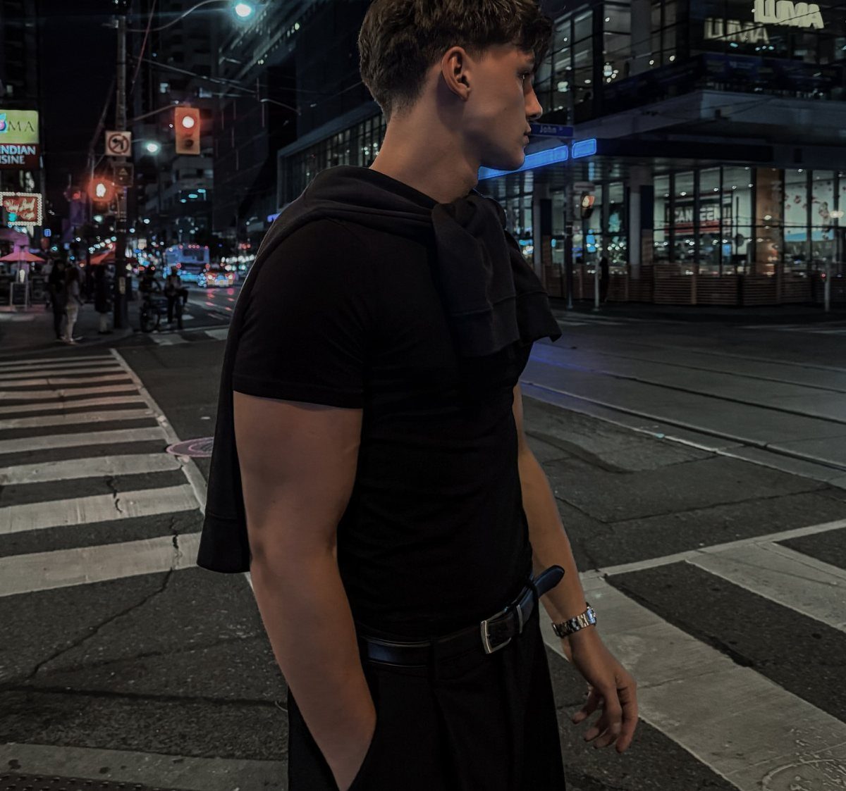 A young man wearing a fitted black t-shirt and trousers standing on a city street at night.