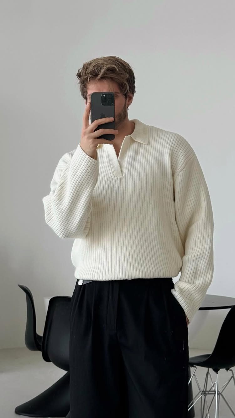 A man in a cream ribbed sweater and black trousers taking a mirror selfie.