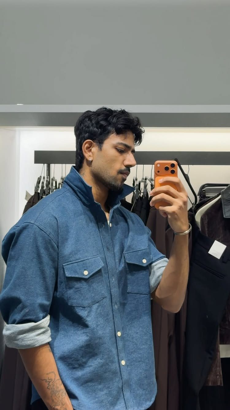 A man in a blue denim shirt taking a mirror selfie in a clothing store.