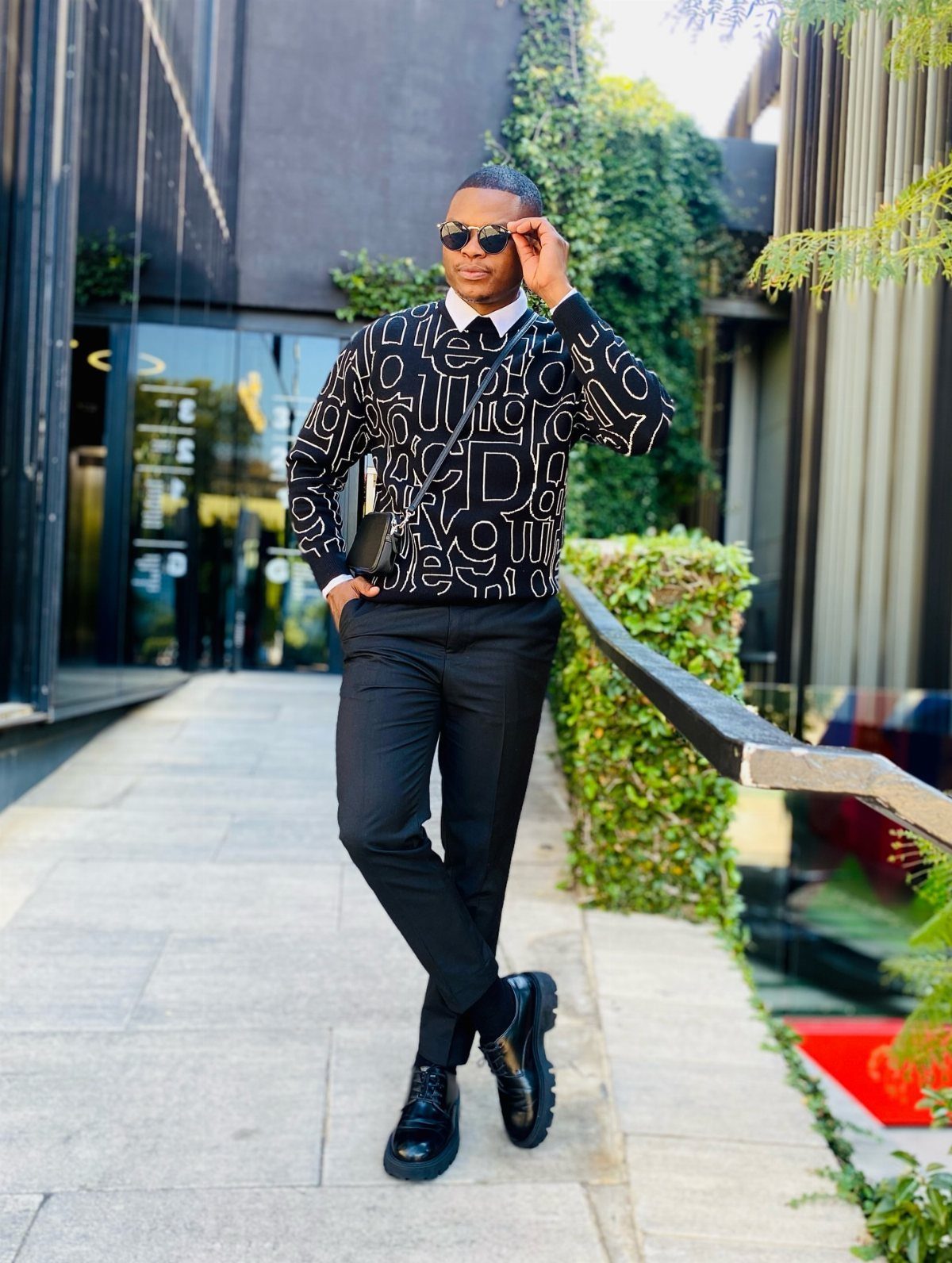 A man in a patterned black shirt and black trousers posing outdoors in sunglasses.