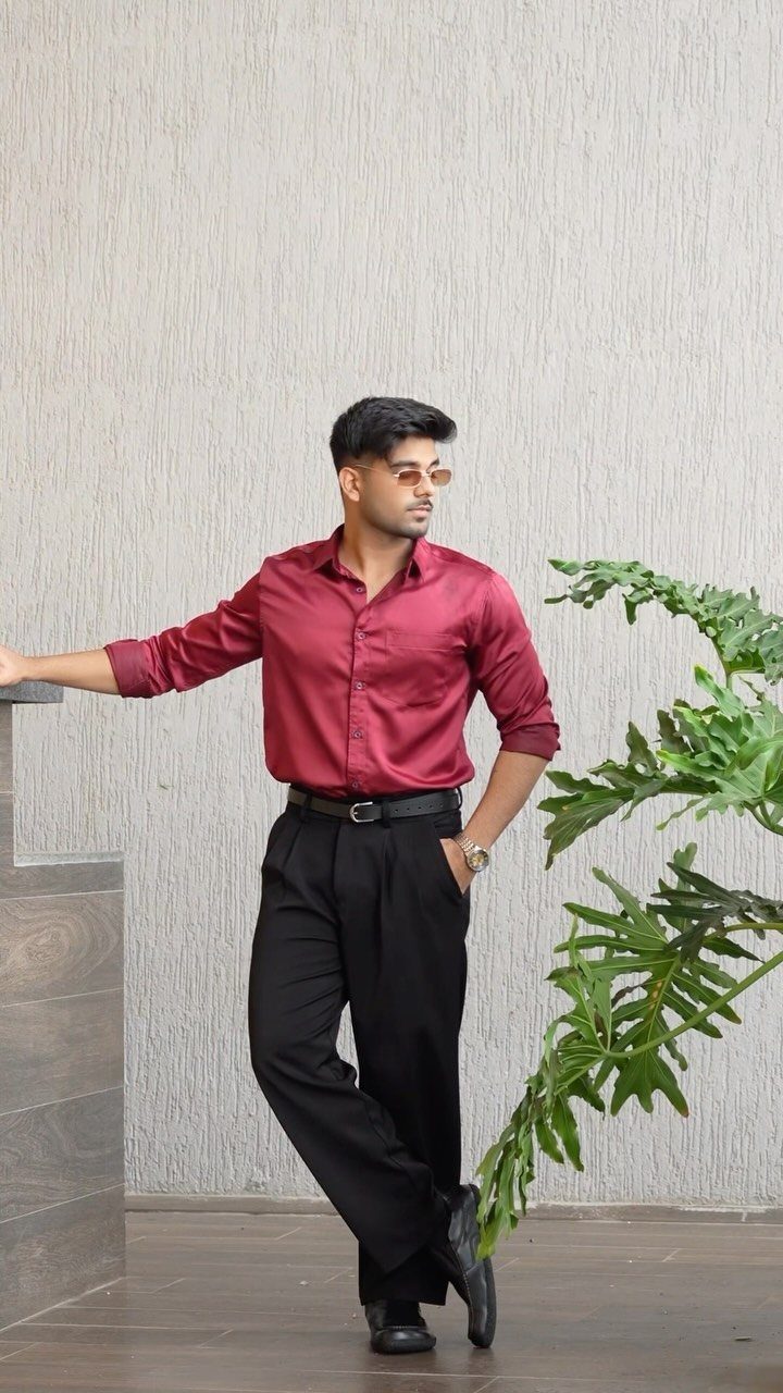 Man in a maroon shirt and black trousers posing against a grey wall.