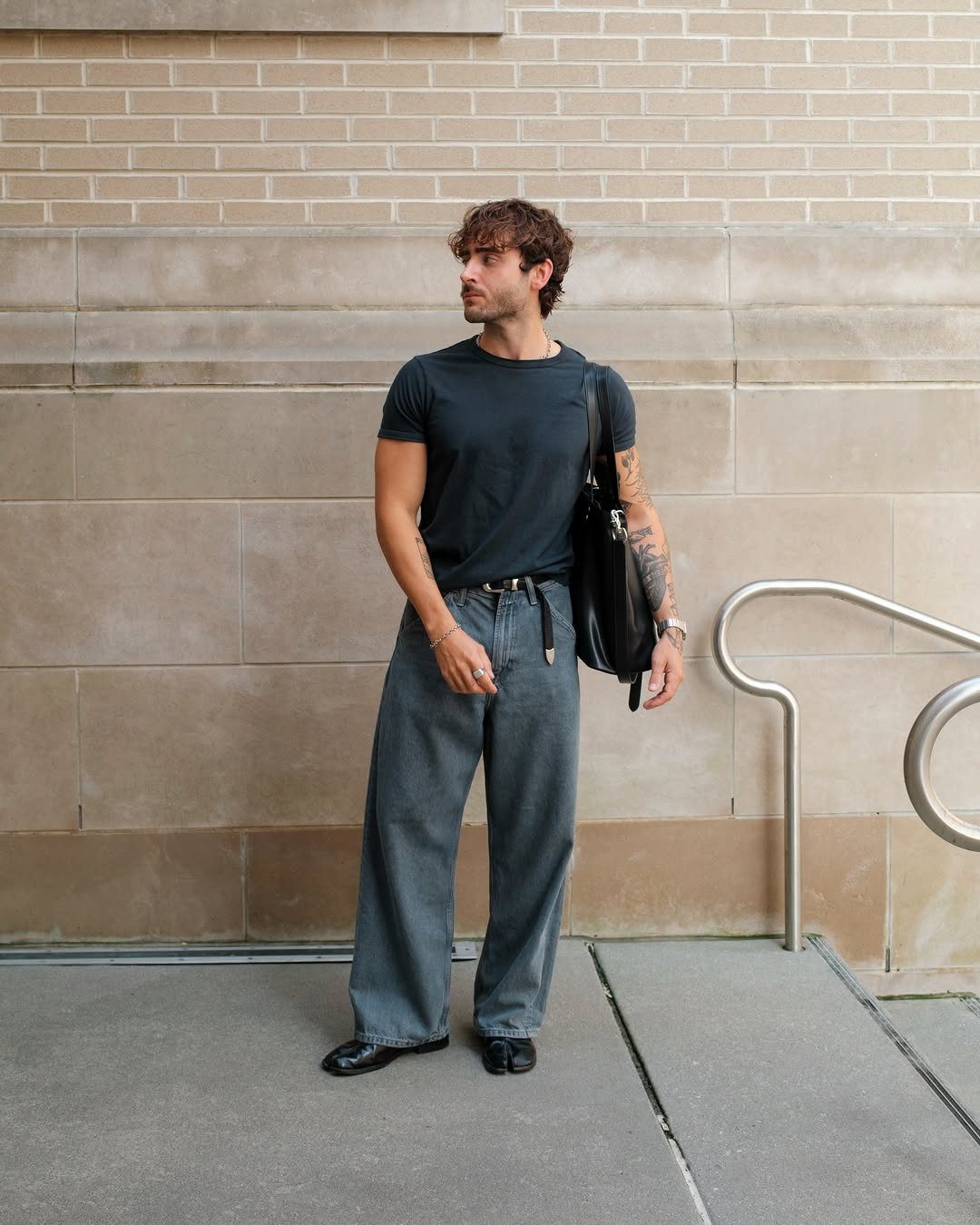 A man wearing a dark t-shirt and wide-leg denim jeans standing against a brick wall.