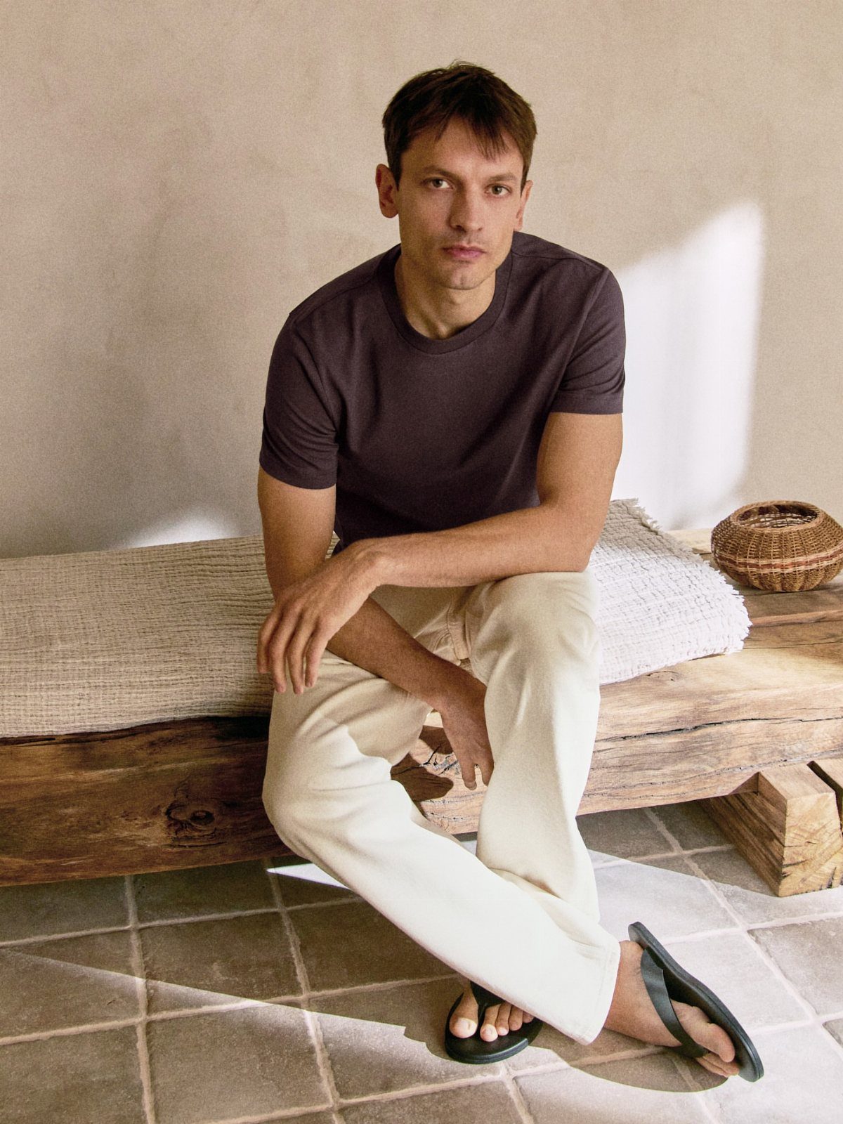 A man sitting on a wooden bench wearing a brown t-shirt and cream trousers.