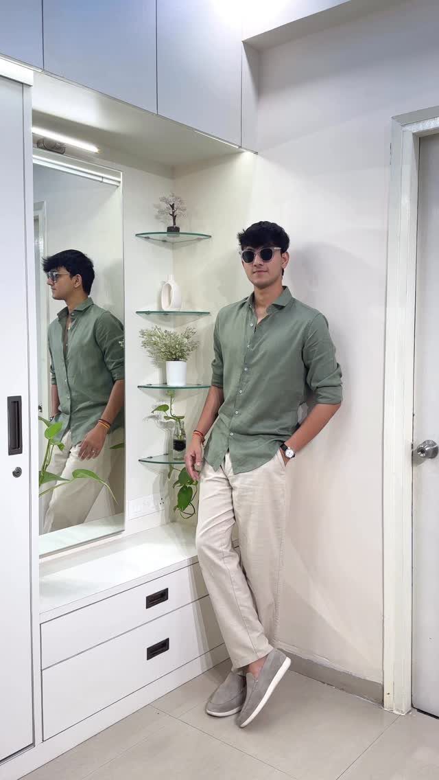 A young man in a green shirt and beige trousers posing in a white room.