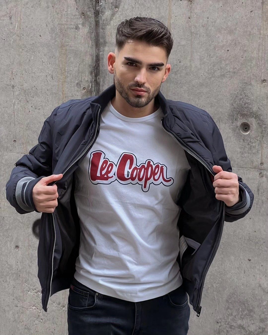 A stylish young man posing in a bomber jacket and graphic t-shirt against a concrete wall.