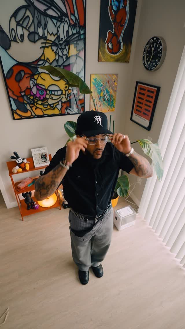 A man in a black shirt and grey trousers posing while adjusting his glasses.
