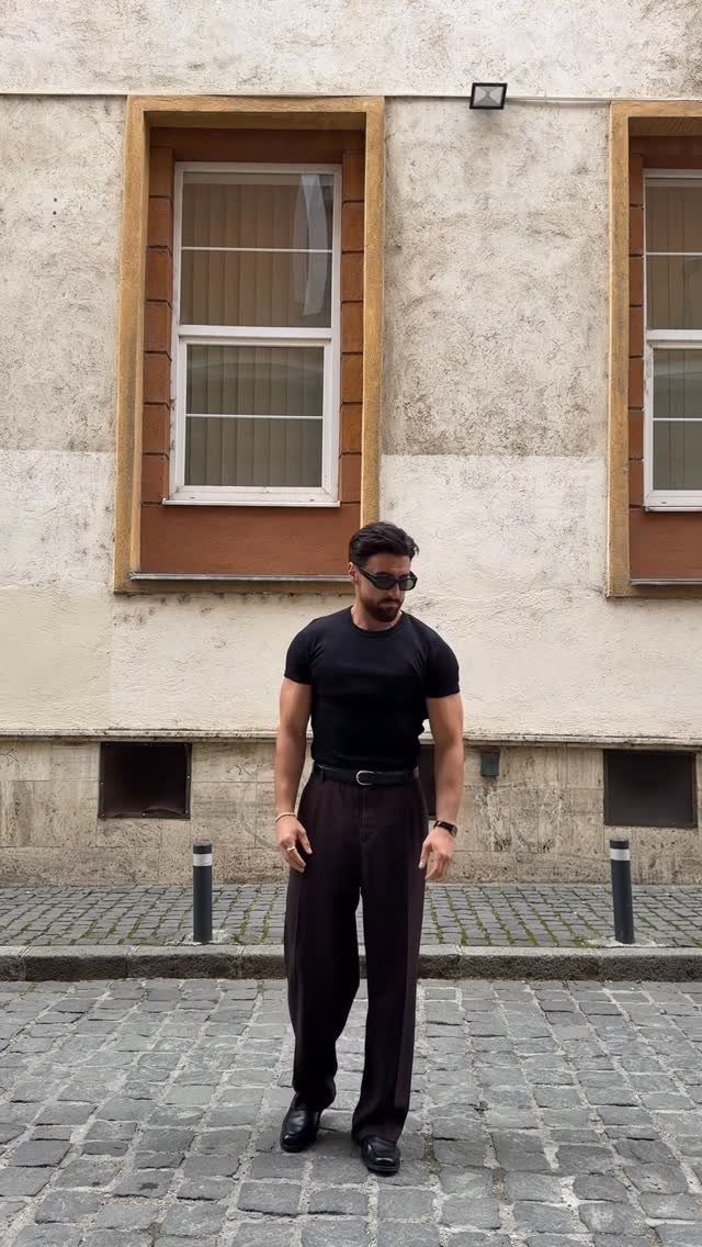 A man in a black t-shirt and brown trousers posing on a cobblestone street.