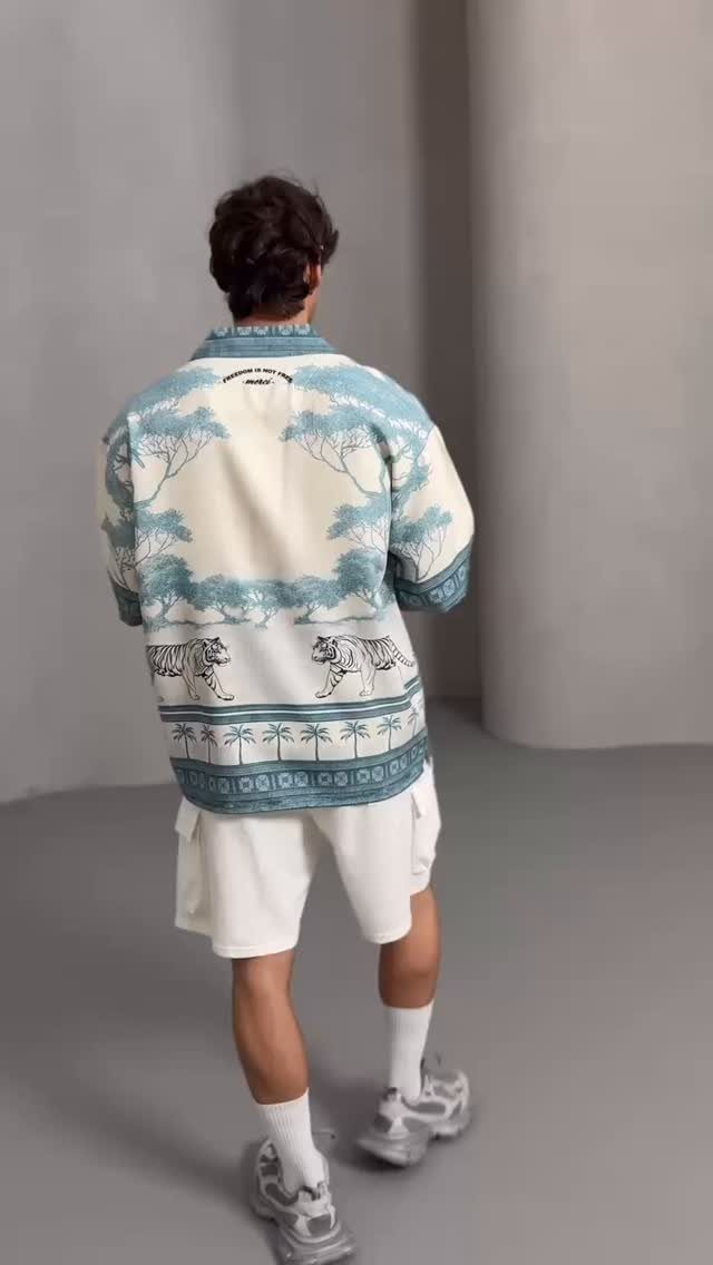 Man wearing a tropical print shirt, white shorts, and sneakers standing in a grey studio.