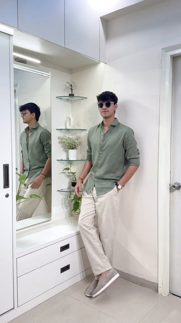 A young man in a green shirt and beige trousers posing against a white wall.