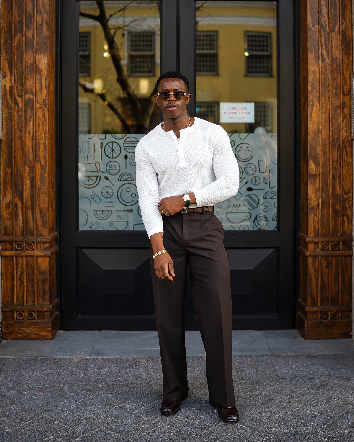 A stylish man in a white henley and brown trousers posing confidently in front of dark doors.