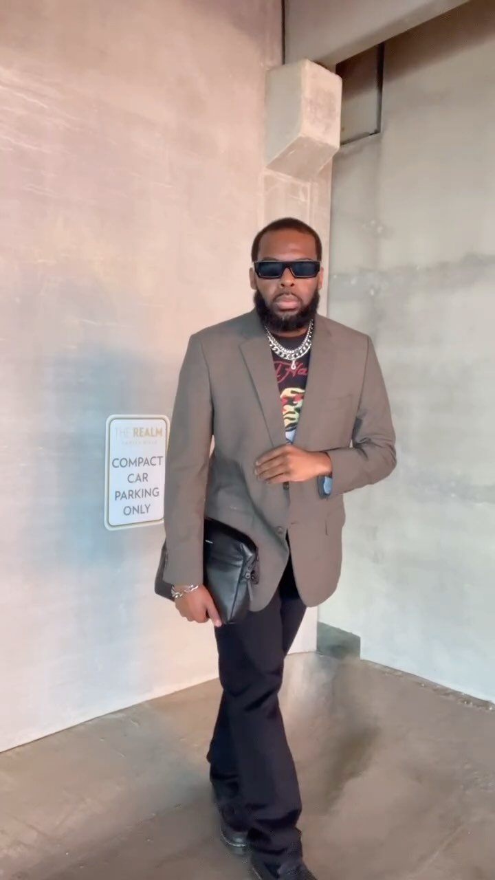 A man posing in a grey blazer, graphic t-shirt, and black trousers against a wall.