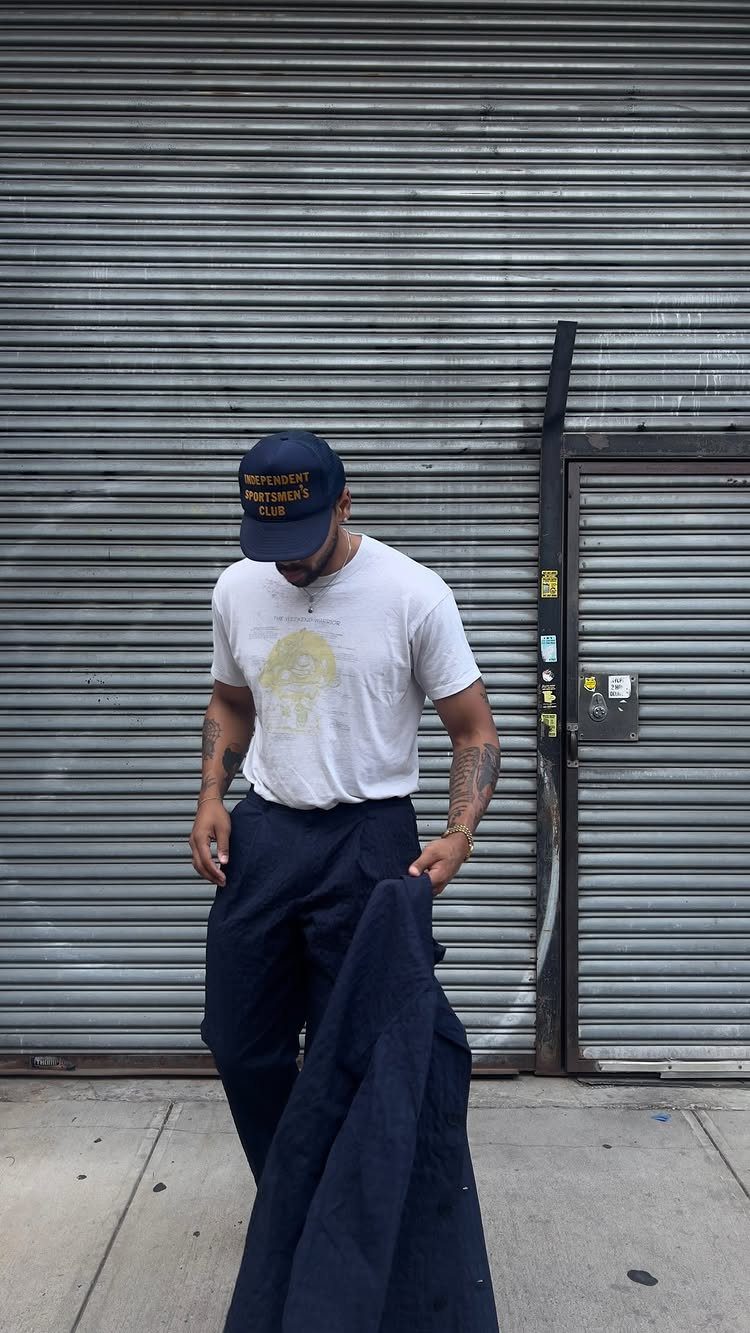 A man wearing a white graphic t-shirt and navy trousers standing before a metal shutter.