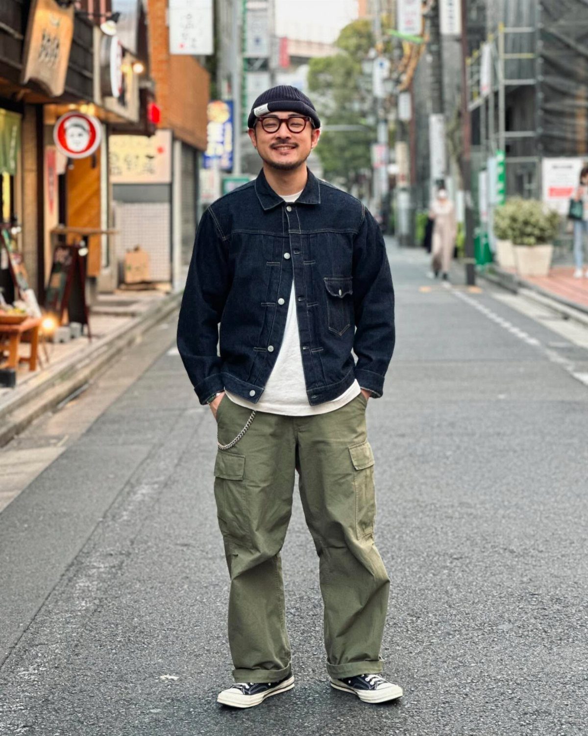 A man wearing a dark denim jacket, white t-shirt, and green cargo pants standing on a street.