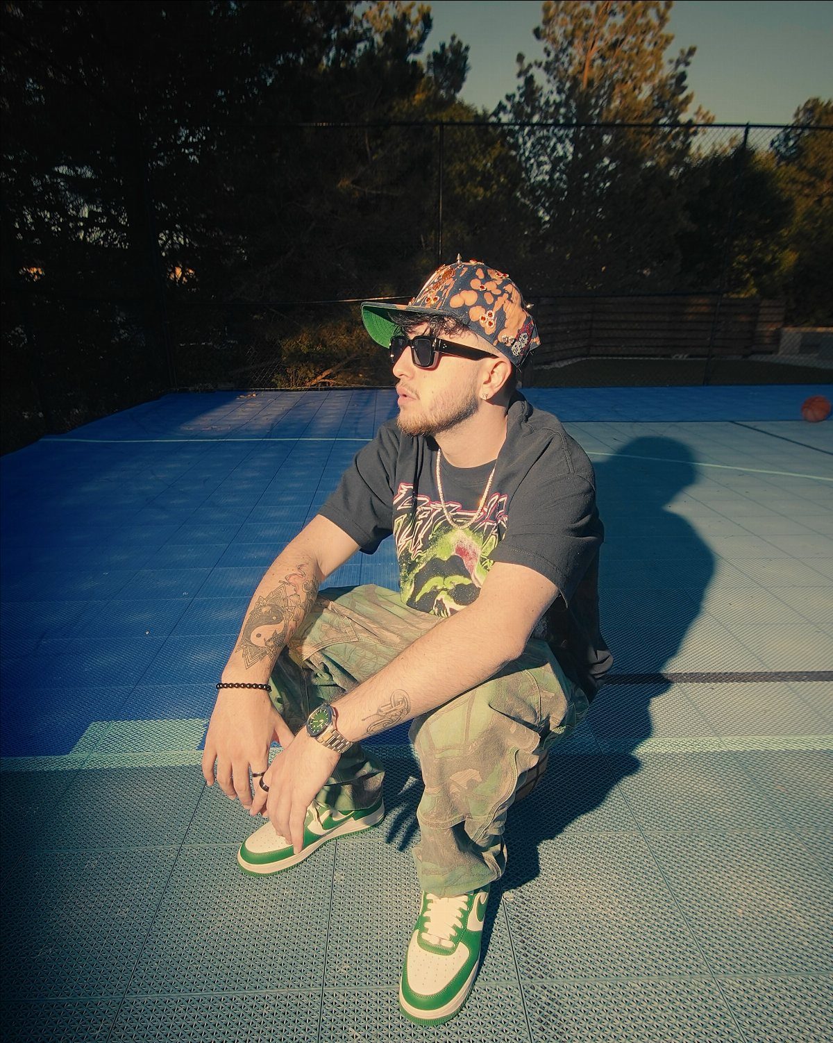 Man in camouflage cargo pants, graphic tee, and green sneakers crouching on a blue court.