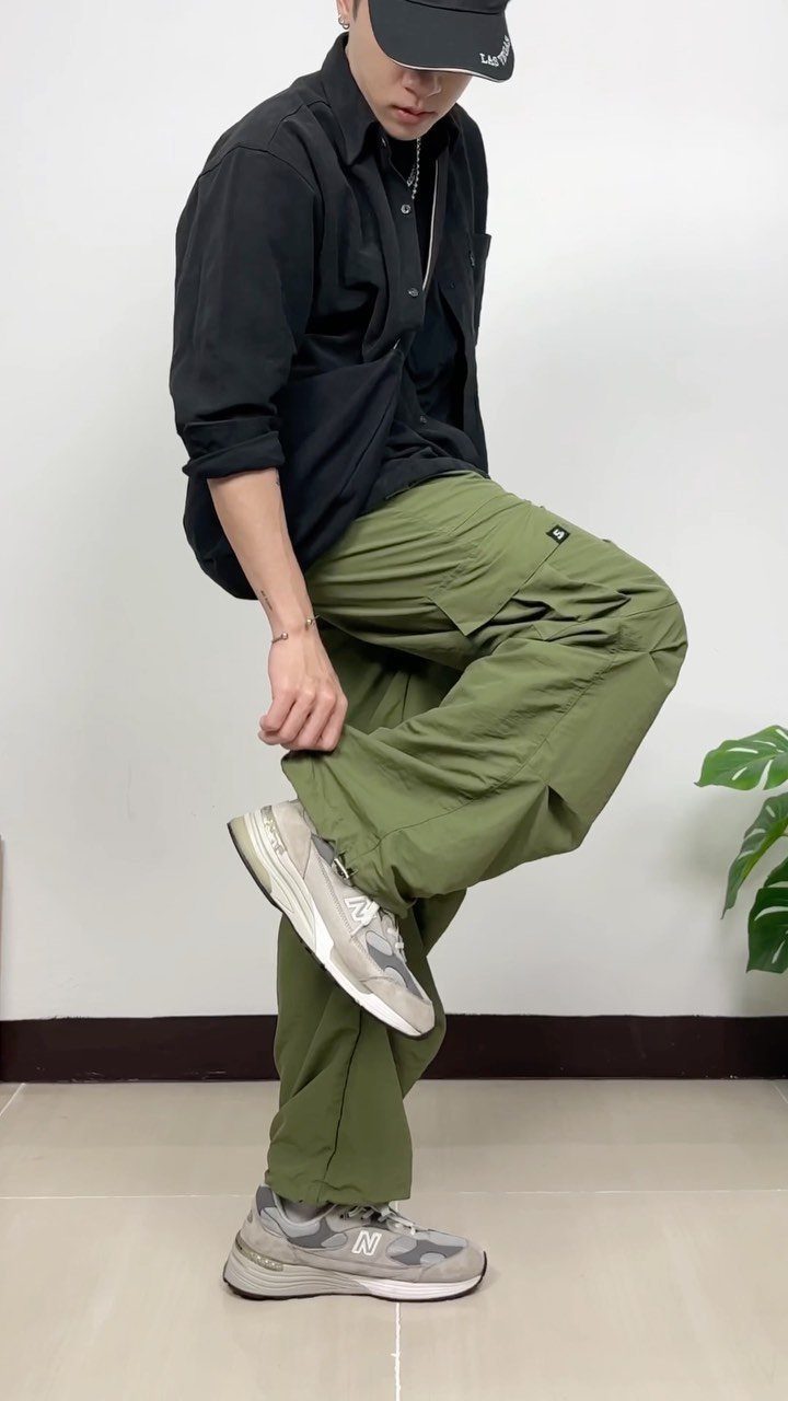 A person wearing green cargo pants, a black shirt, and grey sneakers against a white wall.