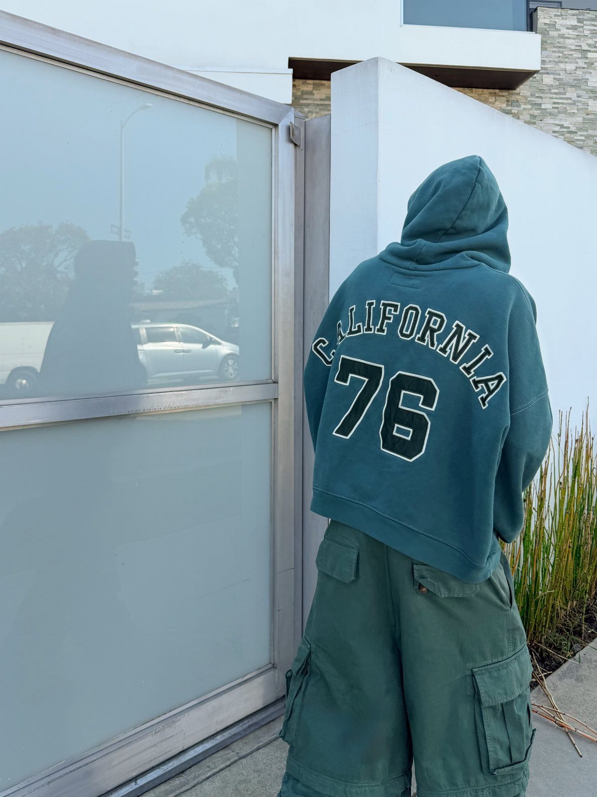 Person wearing a teal California hoodie and green cargo pants standing by a glass window.