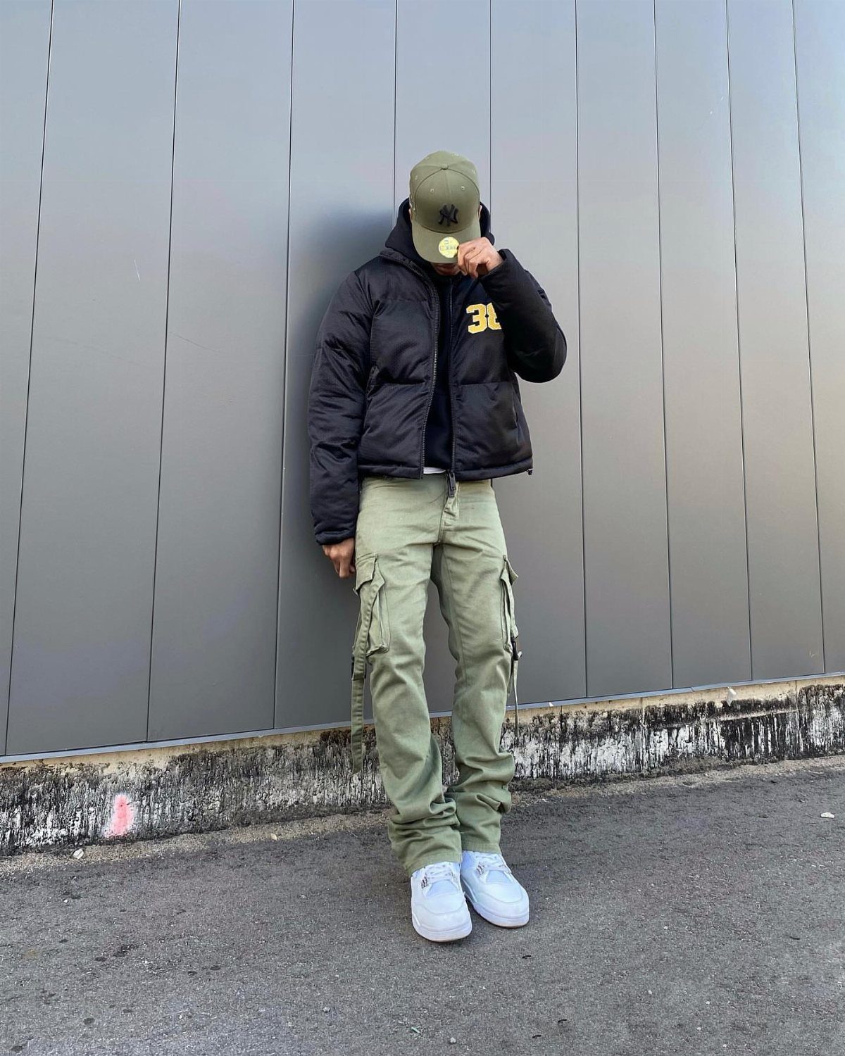 A person wearing a black puffer jacket and olive green cargo pants standing against a grey wall.