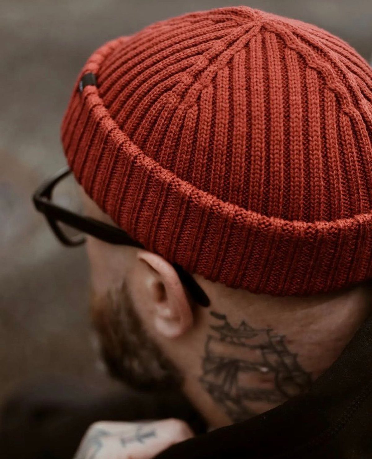 A man wearing a burnt orange ribbed knit beanie hat with a ship tattoo on his neck.