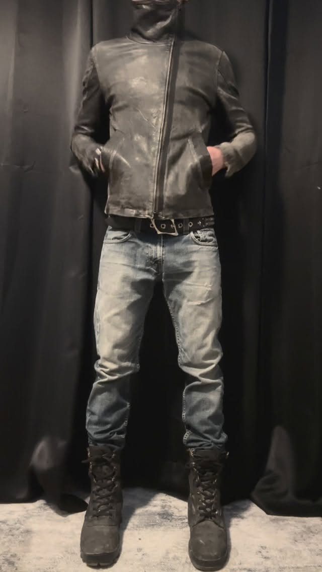 Man wearing a distressed grey jacket, blue jeans, and black combat boots against a black background.