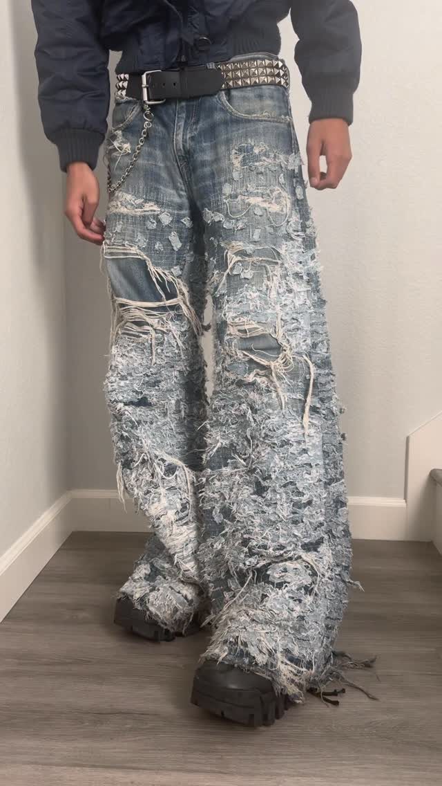 Extremely distressed wide-leg blue denim jeans with heavy fraying and large holes worn by a person.