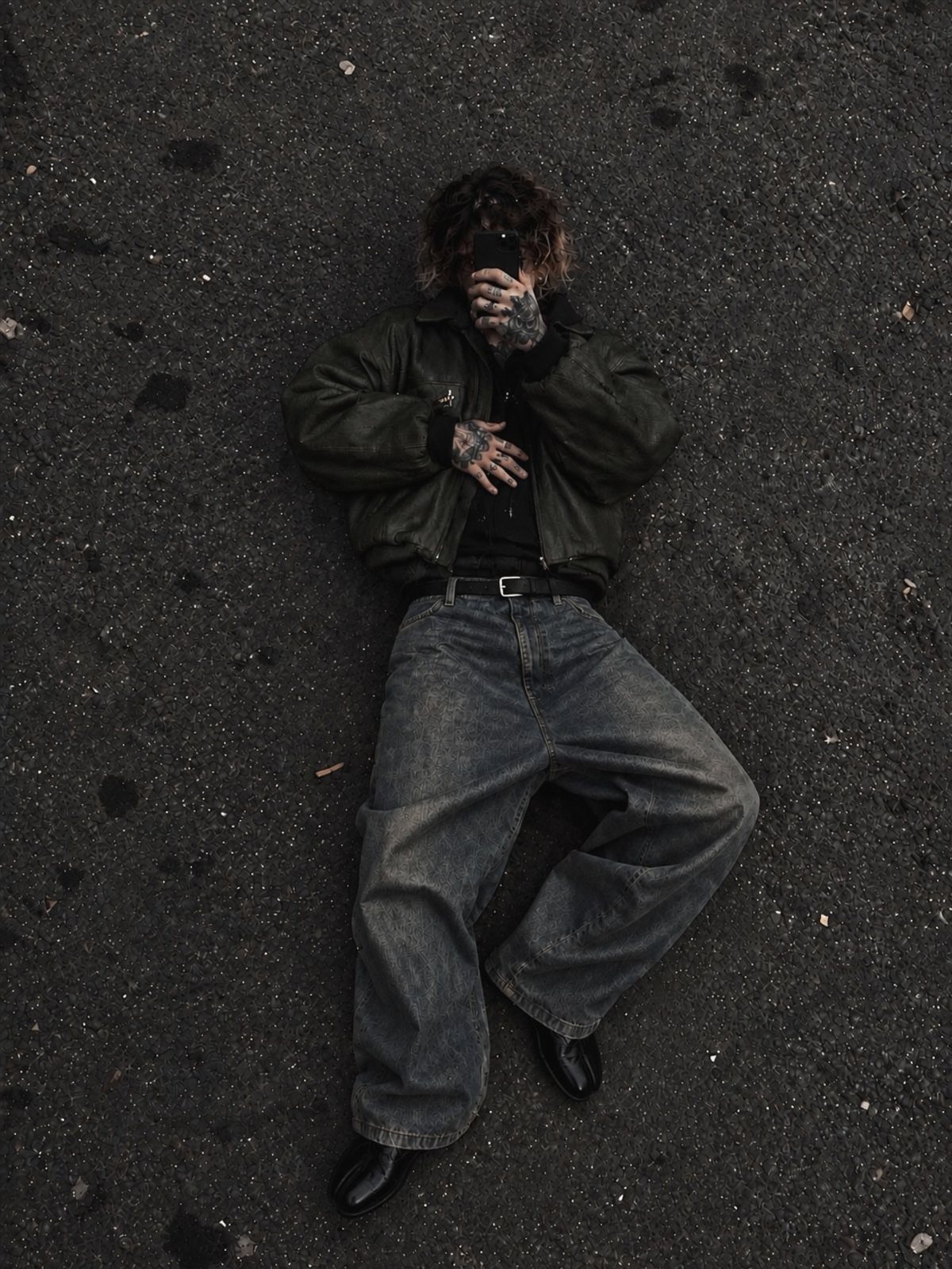 A person in baggy denim and a bomber jacket lying on dark asphalt.