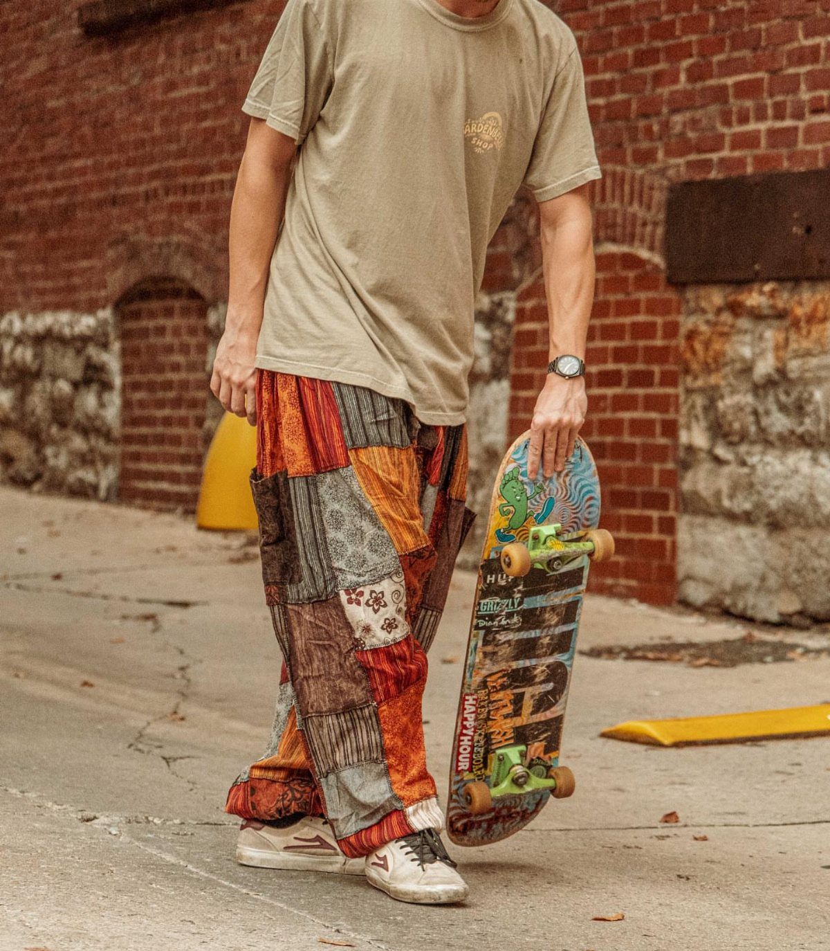 A person in patchwork trousers walks while carrying a skateboard next to a brick wall.