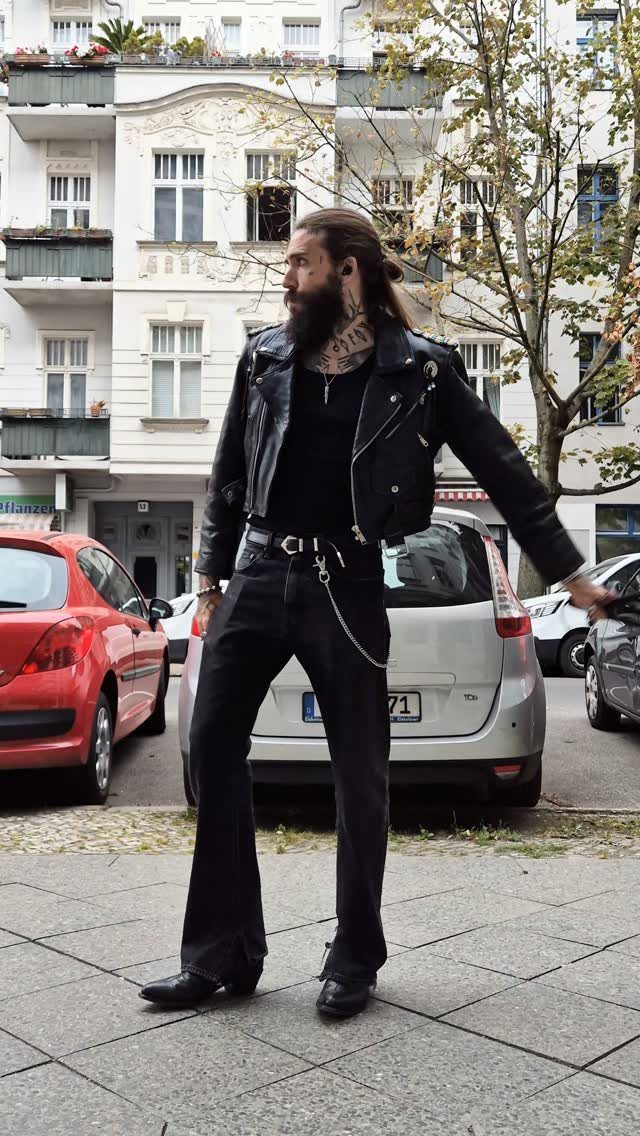 Man with a beard and tattoos wearing a black leather jacket and dark clothing on a city street.