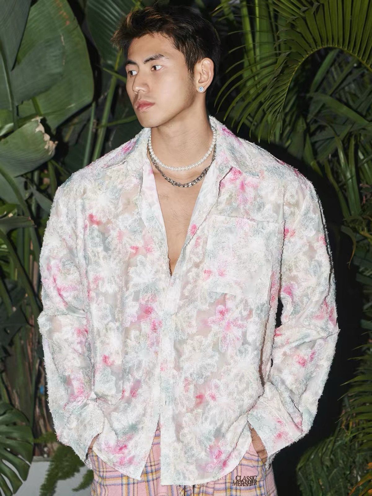 A young man wearing a light floral patterned shirt and pearl necklaces amidst tropical greenery.