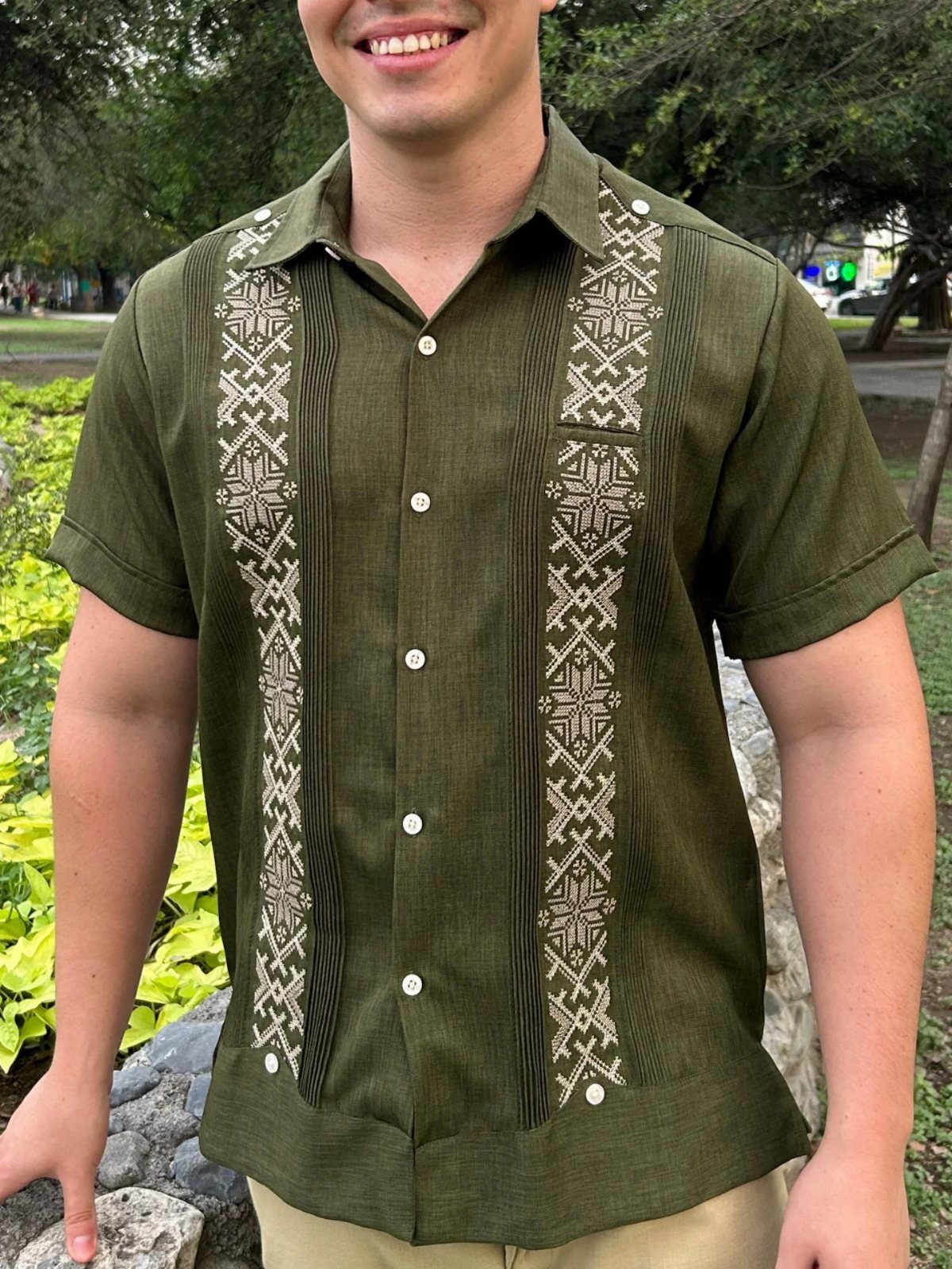 A man smiling while wearing an olive green embroidered guayabera shirt outdoors.