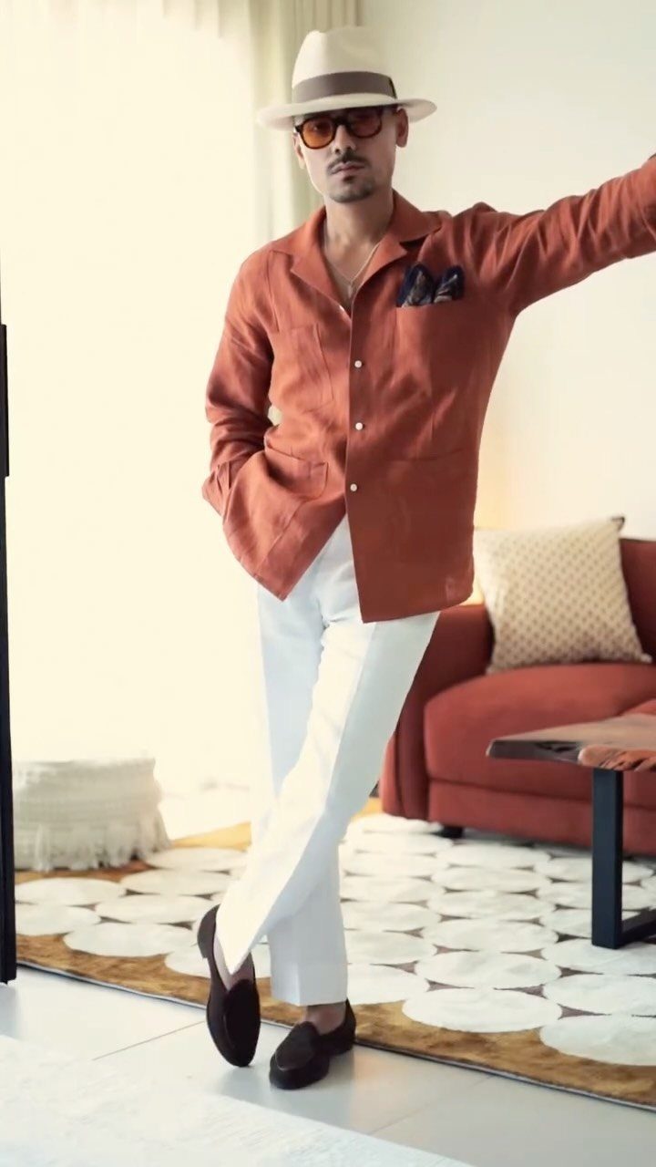 Man in terracotta linen shirt, white pants, and fedora posing in a bright room.