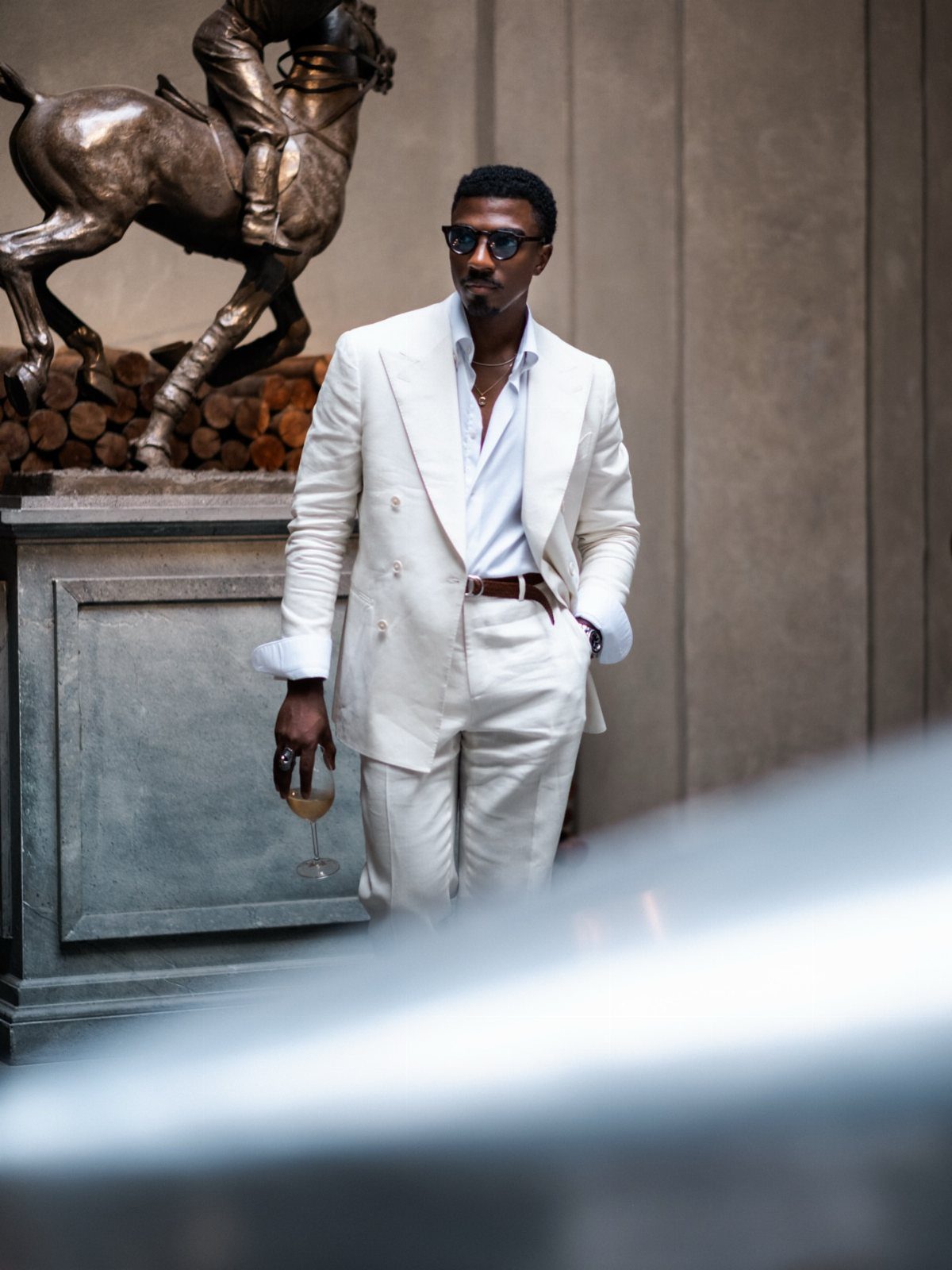 A man in a white double-breasted suit and sunglasses standing by a bronze equestrian statue.