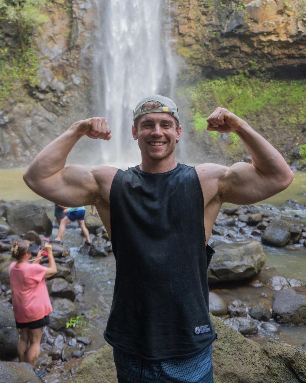Muscular man in black athletic tank top flexing his arms in front of a waterfall.