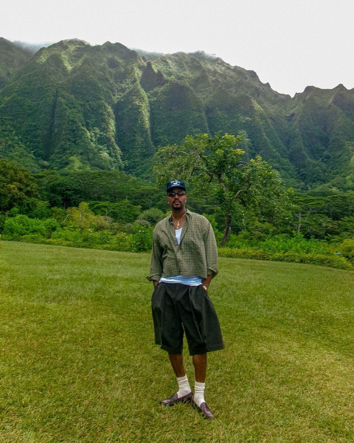 A man wearing dark knee-length shorts and a patterned shirt standing in a grassy field.