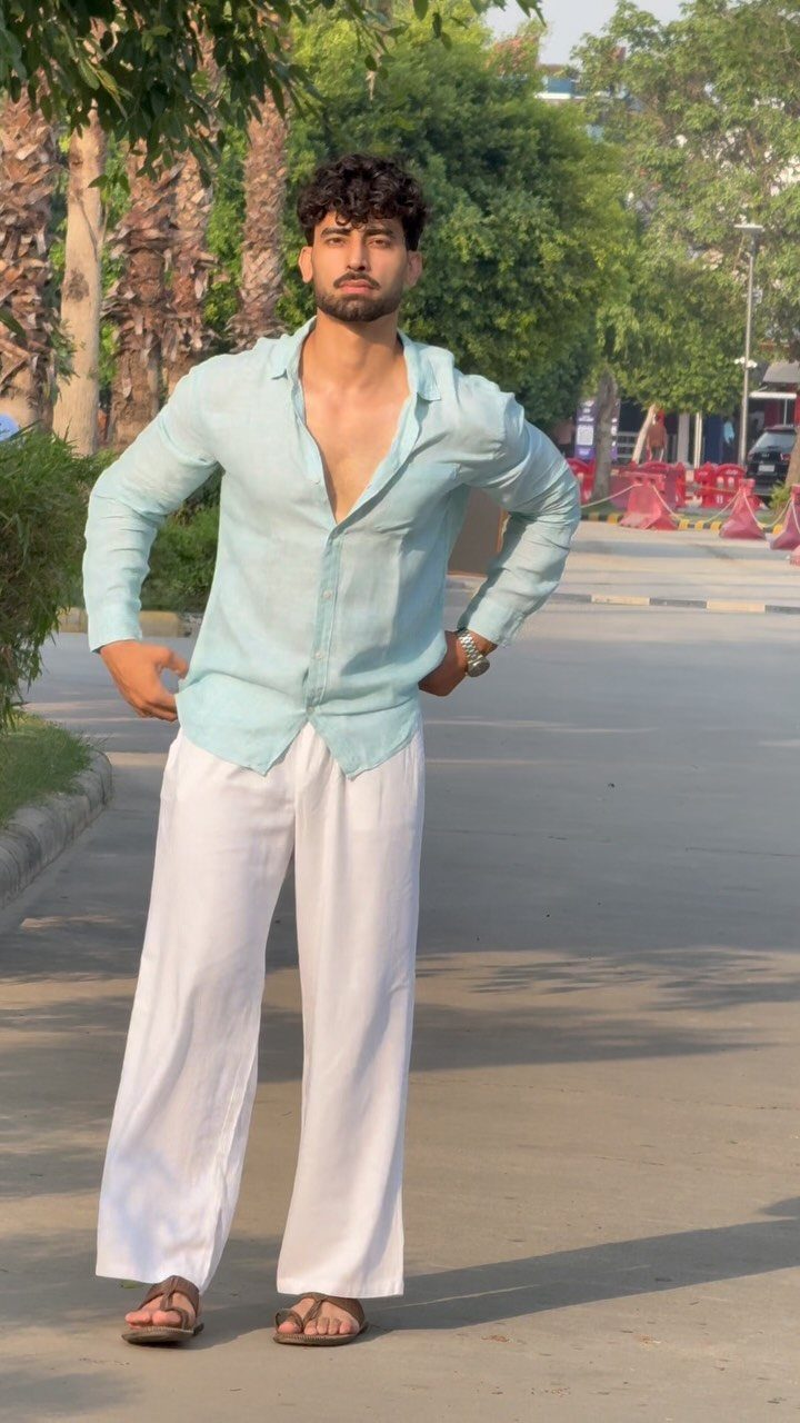 A man posing in a light blue shirt and wide-leg white linen pants.