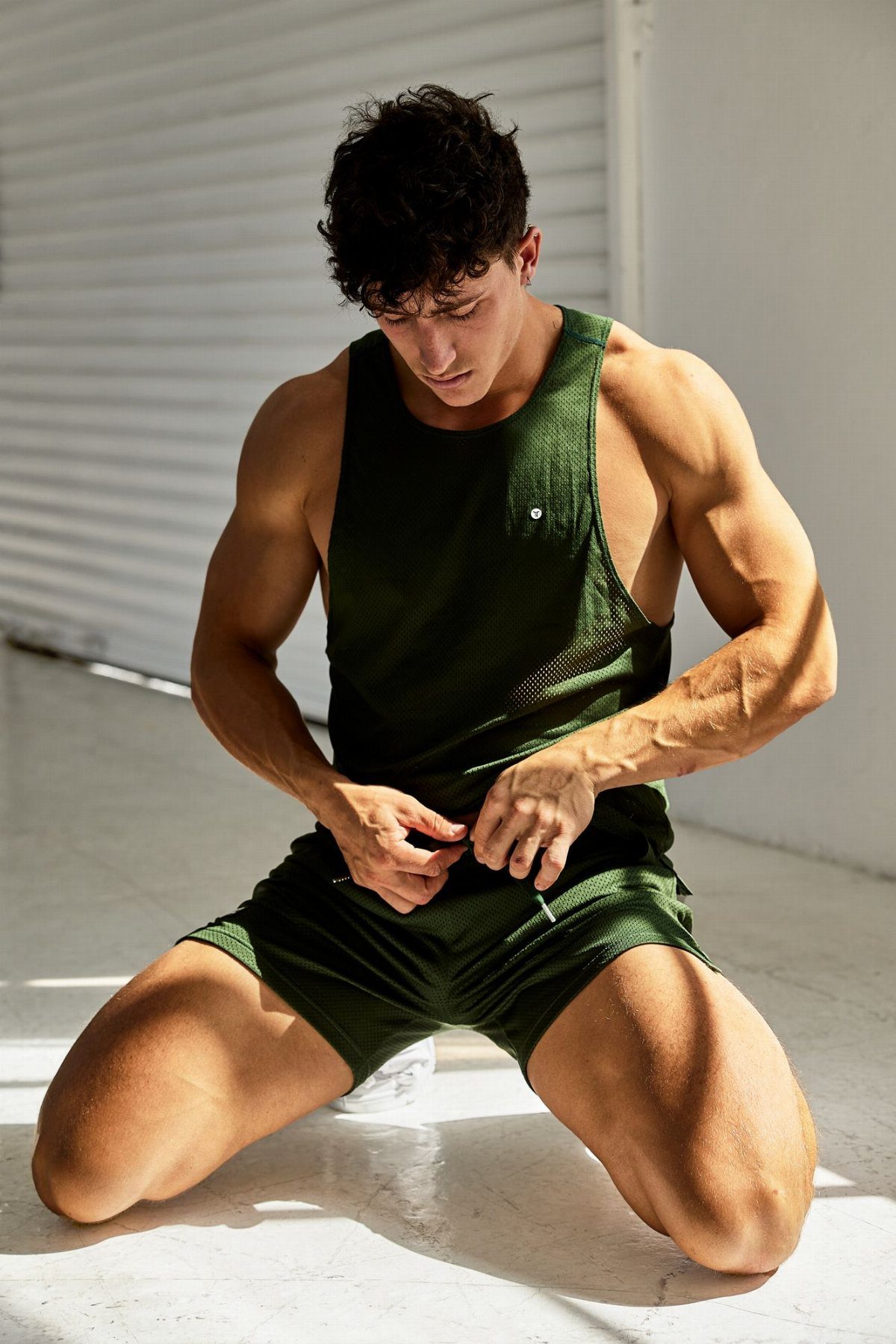 A muscular man wearing a dark green mesh athletic tank top and shorts.