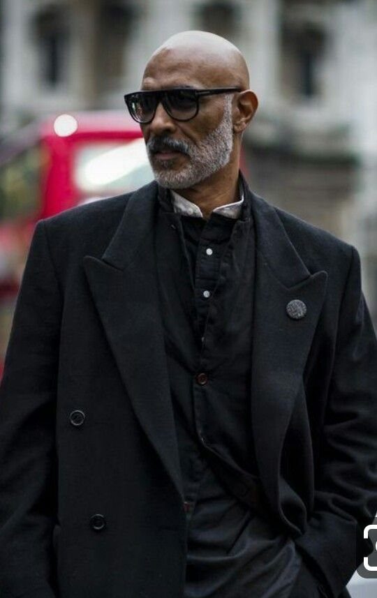 A man with a grey beard wearing black glasses and layered black clothing outdoors.