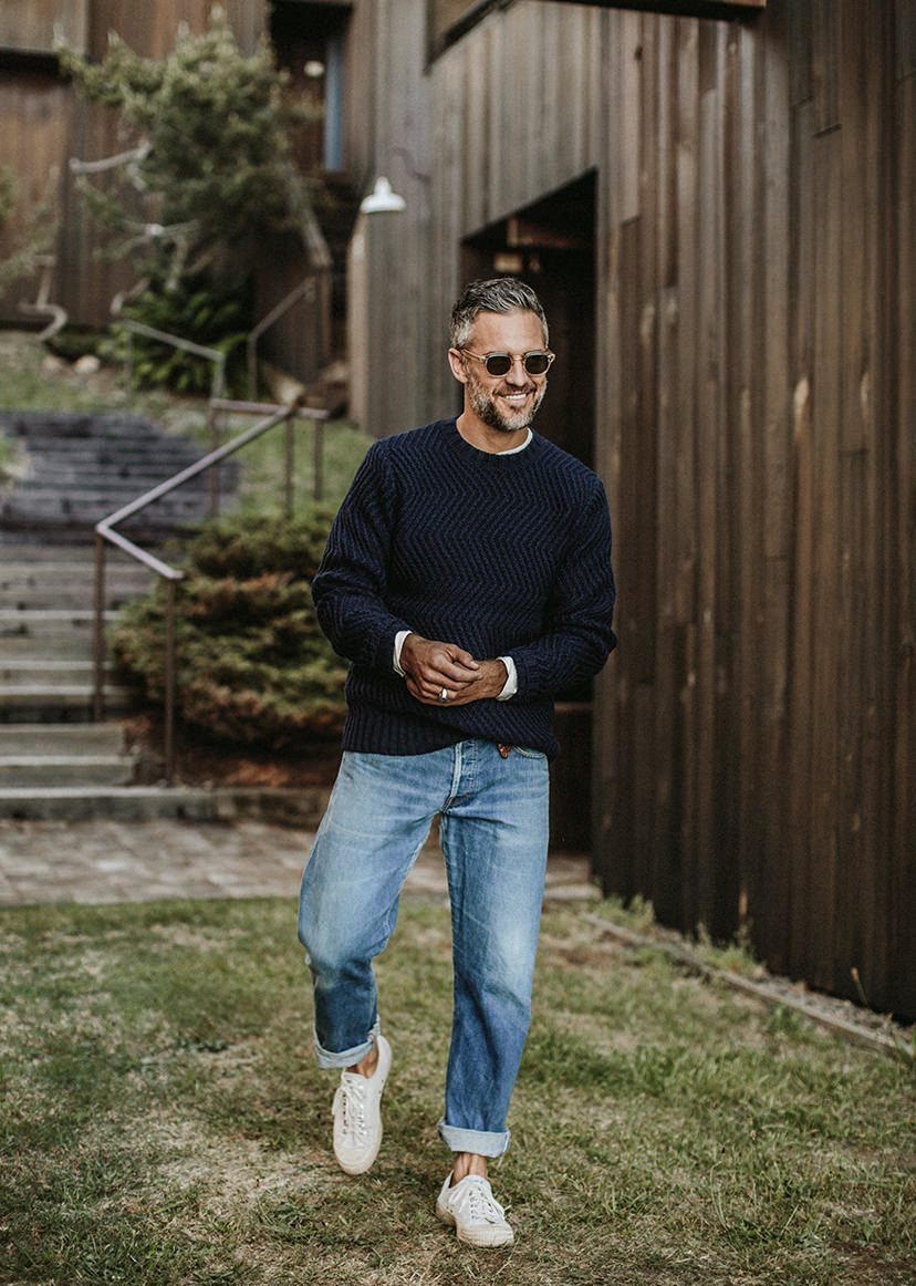 A smiling man in a navy sweater and blue jeans walking past a wooden building.