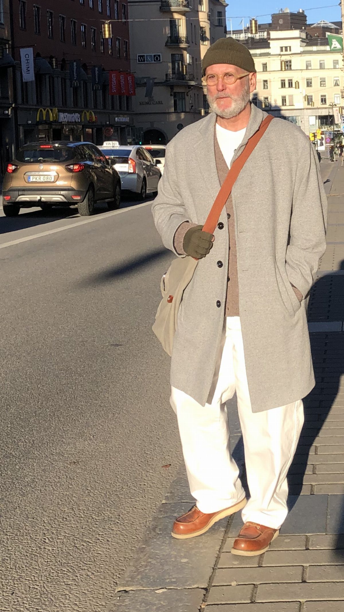 An older man posing on a city street wearing an oversized grey coat and white trousers.